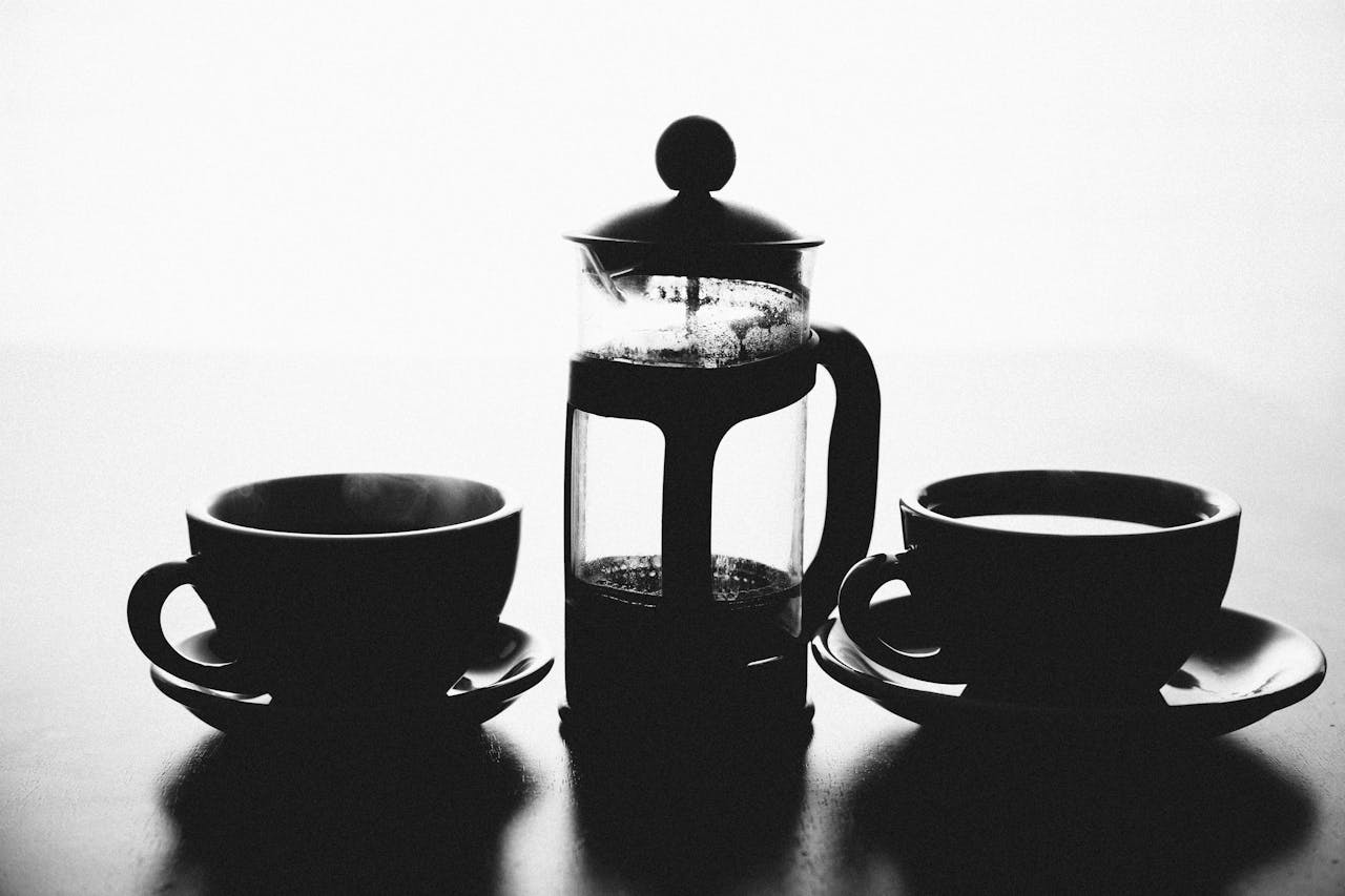 Monochrome image of a French press and coffee cups for a classic coffee experience.